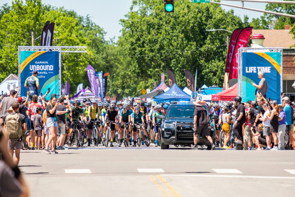 A large volume of riders waits at the Unbound 2022 XL start line behind a police escort that is about to start rolling out. 