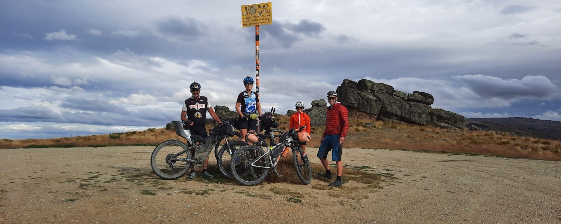A group of cyclists stands near a road sign. All are posed on or near their bikes and are looking toward the camera with a smile.