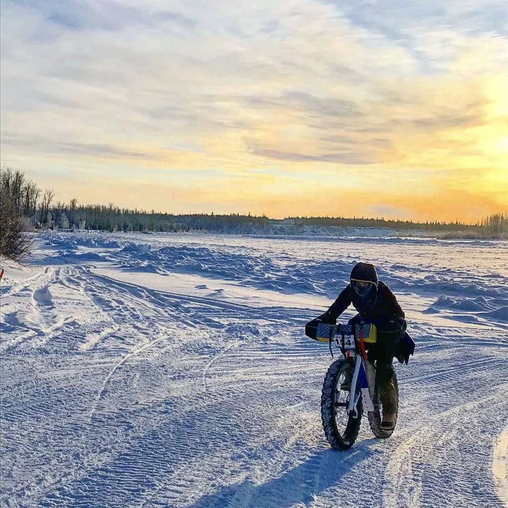 A cyclist rides her Otso Cycles Voytek fat bike across hard-packed Alaskan snow as the sun sets behind her.