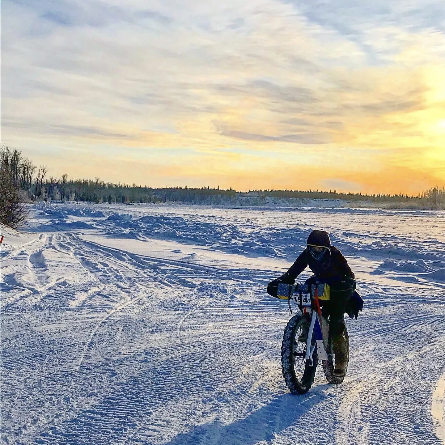 A cyclist rides hard packed snow in Alaska. There is a wide expanse of white snow. The cyclist has her head down and is powering through on her Otso Cycles Voytek fat bike.