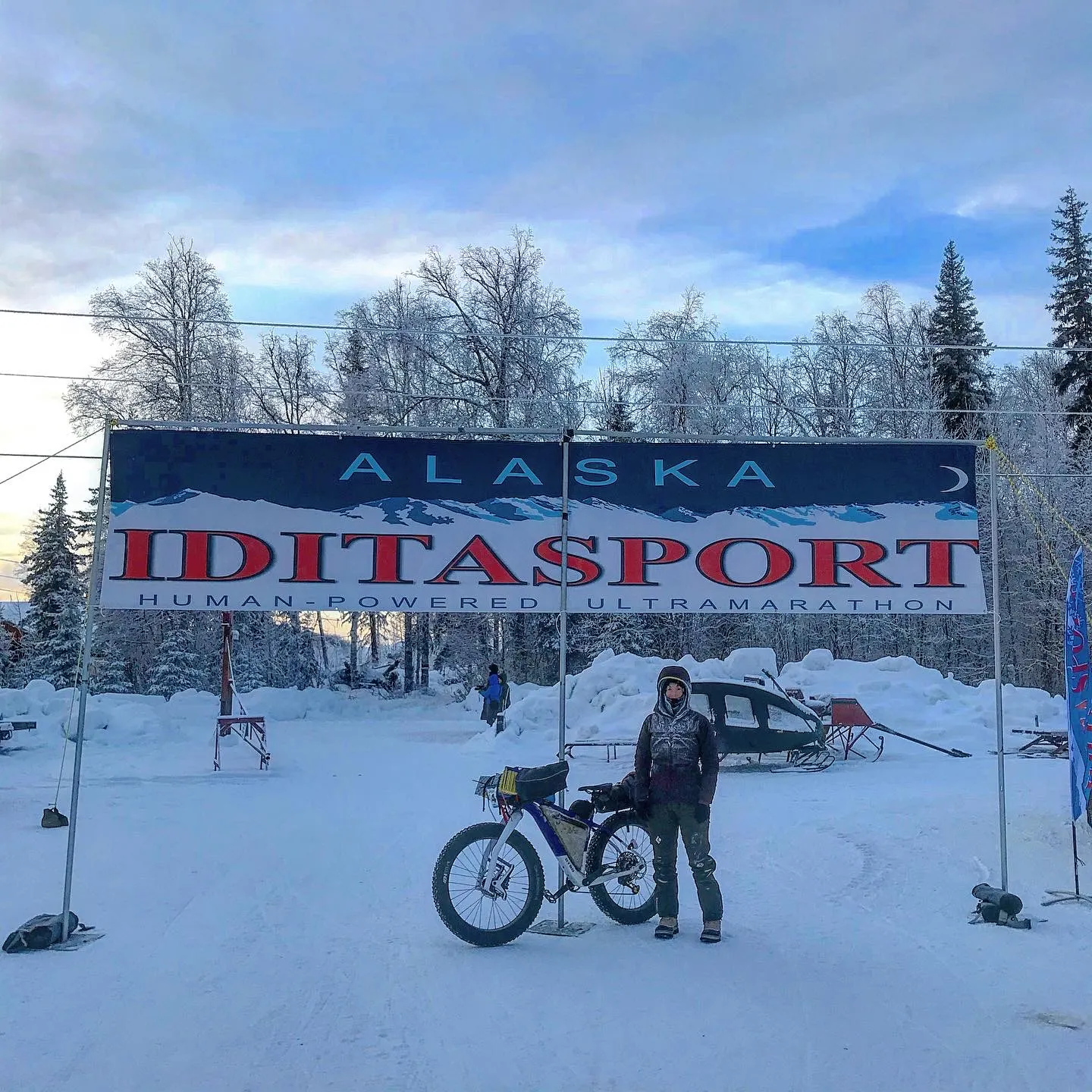 A cyclist stands underneath a sign that reads Alaska Iditasport Human-Powered Ultramarathon. The cyclist is standing next to her Otso Cycles Voytek fat bike.