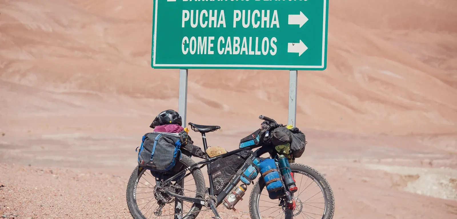 An Otso Cycles Voytek fat bike leans against a street sign in the Andes Mountains. The hills behind are covered in rocks and sand. The sign reads Barrancas Blancas, Pucha Pucha, and Come Caballos.