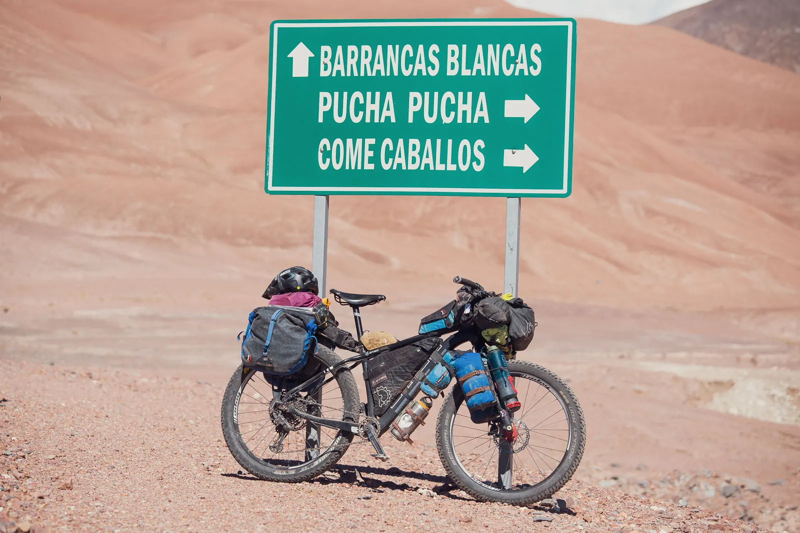 An Otso Cycles Voytek fat bike leans against a street sign in the Andes Mountains. The hills behind are covered in rocks and sand. The sign reads Barrancas Blancas, Pucha Pucha, and Come Caballos.