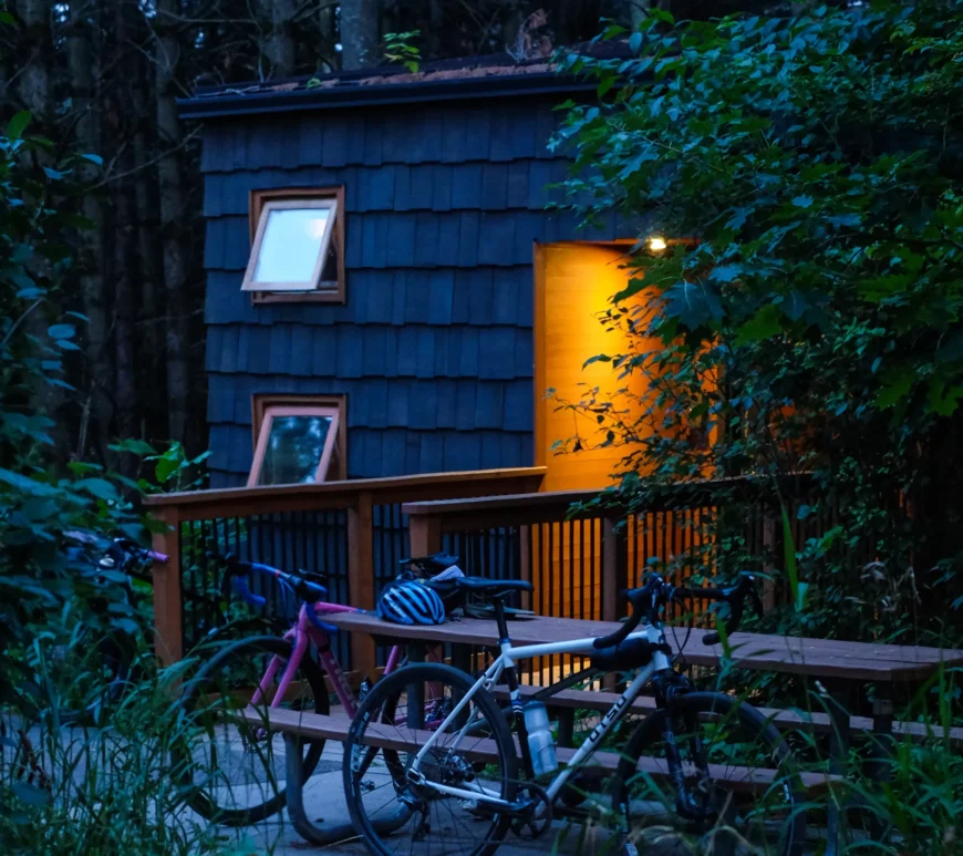 A photo of a cabin after twilight. The sun has descended and it is dark around the cabin. Two bikes lean against a picnic table outside of the cabin, and warm orange lights shine out from the cabin's doorway.
