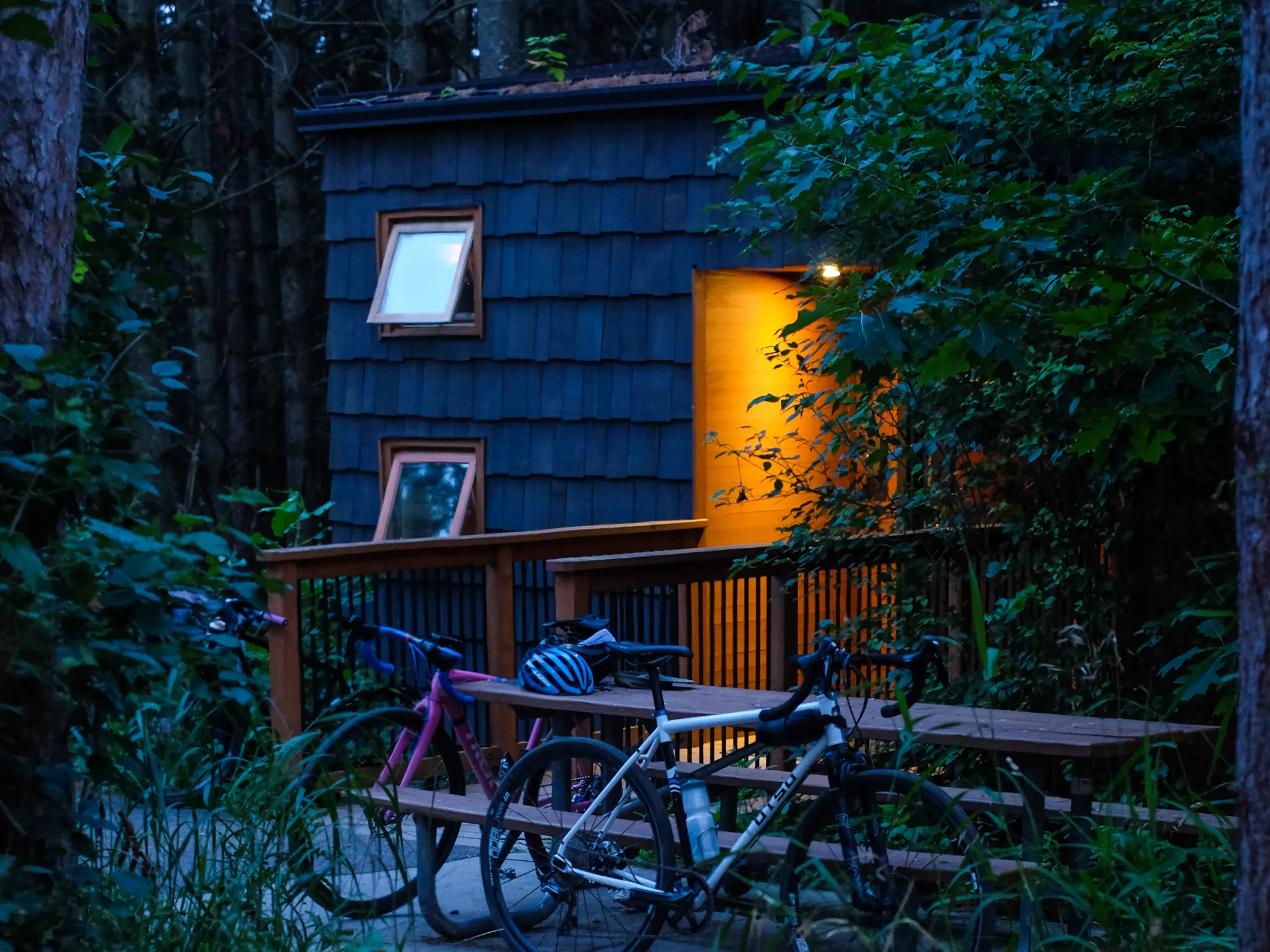 A photo of a cabin after twilight. The sun has descended and it is dark around the cabin. Two bikes lean against a picnic table outside of the cabin, and warm orange lights shine out from the cabin's doorway.