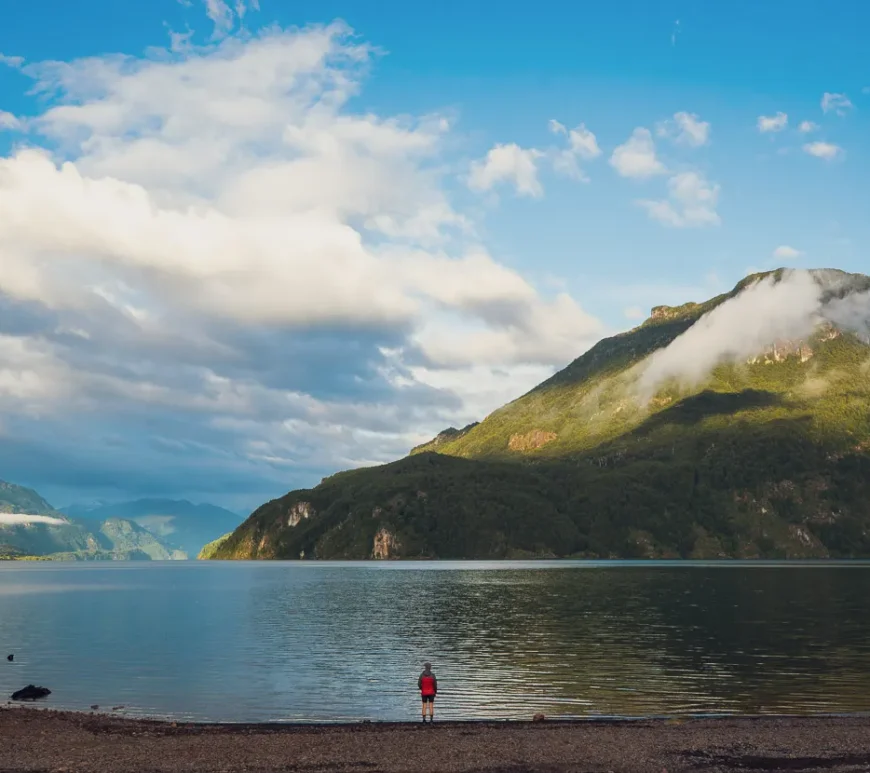 A wide landscape image that shows a clear lake. Behind the lake a mountain extends up towards the clouds. A lone individual is standing at the edge of the lake. Because of the image's perspective, the individual looks very small.