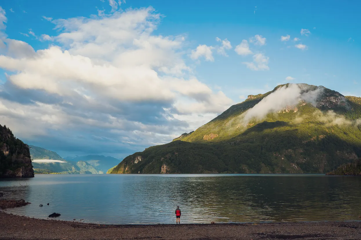 A wide landscape image that shows a clear lake. Behind the lake a mountain extends up towards the clouds. A lone individual is standing at the edge of the lake. Because of the image's perspective, the individual looks very small.