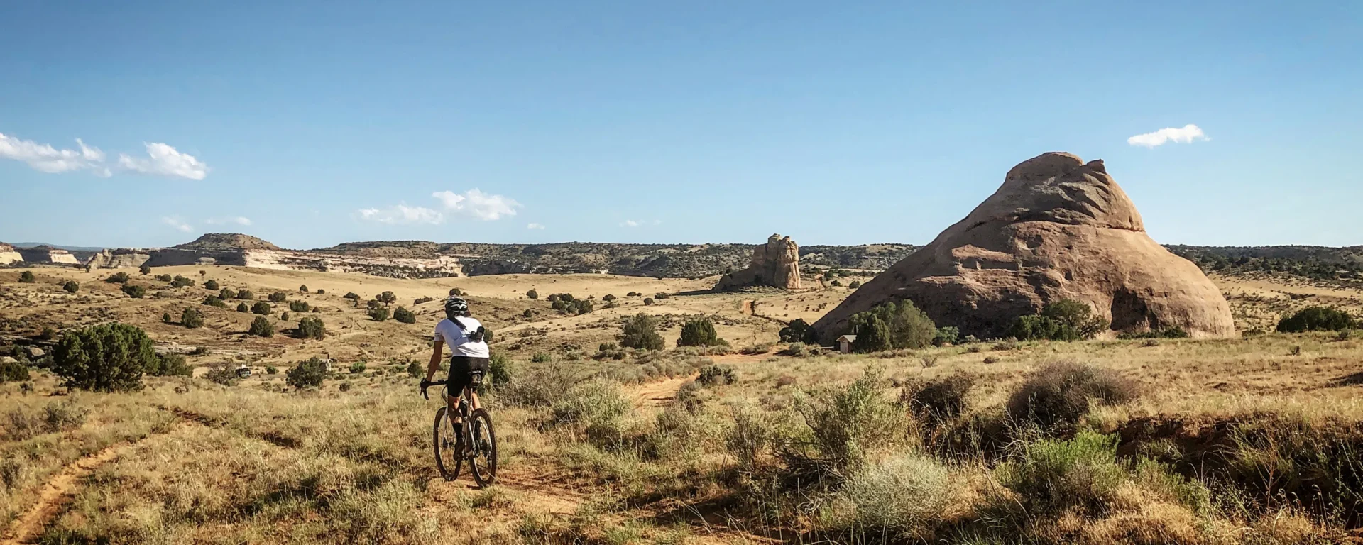 A cyclist rides along a desert gravel path. Rock formations surround the landscape. The sky is a bright blue, and the sun is shining.
