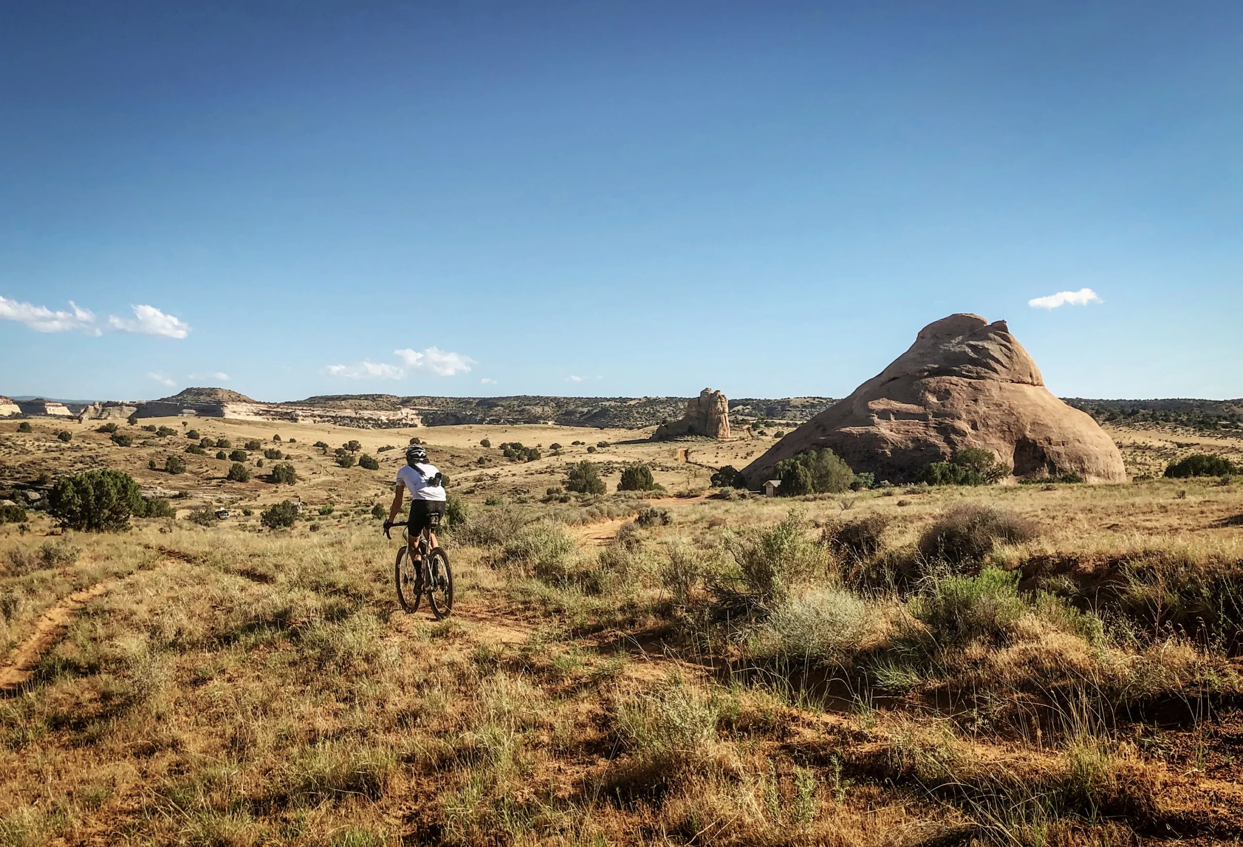 A cyclist rides along a desert gravel path on his Otso Cycles Waheela C. Rock formations surround the landscape. The sky is a bright blue, and the sun is shining.