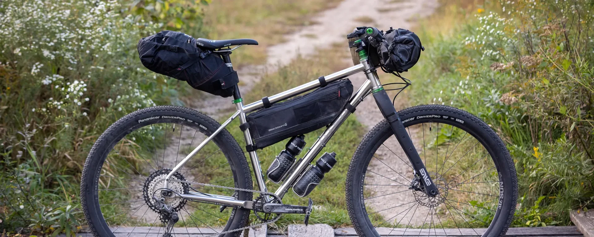 An Otso Cycles Fenrir Stainless positioned on a wooden bridge for photographs. The bike has green Wolf Tooth Components and a suite of bikepacking bags.