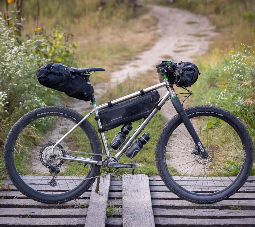An Otso Cycles Fenrir Stainless positioned on a wooden bridge for photographs. The bike has green Wolf Tooth Components and a suite of bikepacking bags.