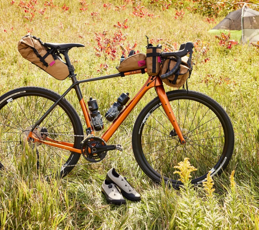 An Otso Cycles Waheela C is posed in a field. The bike is part of a large giveaway. The bike is outfitted with bikepacking bags from Swift Industries. In front of the bike are a pair of riding shoes from QUOC.